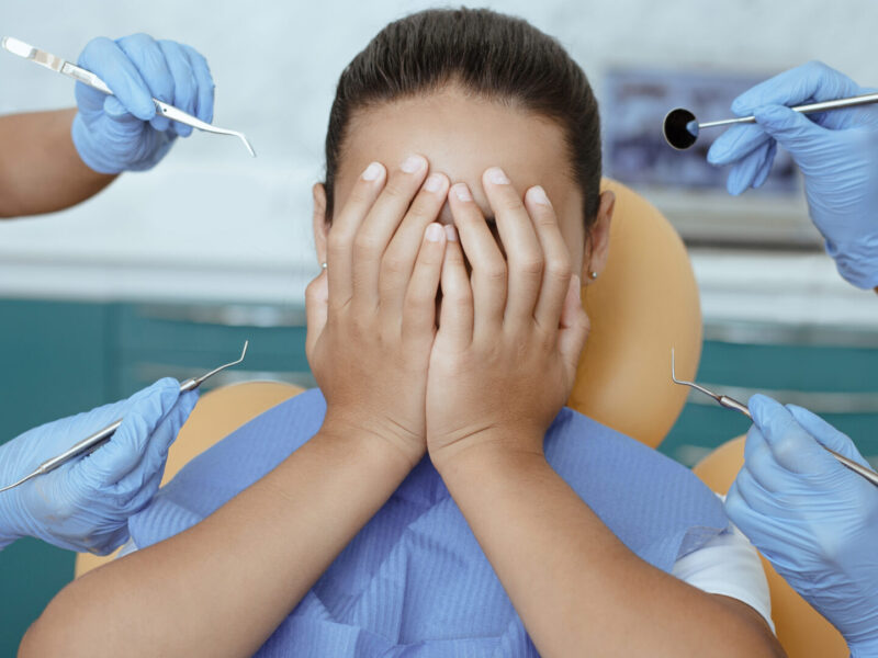 Multiracial doctors in rubber gloves scare girl with medical instruments. Mixed race little girl patient with napkin on chest in dental chair covers face with hands, close up, cropped, cropped